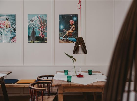 Ein gemütlicher Innenraum mit Holztischen und Stühlen. An der Wand hängen farbenfrohe Kunstwerke.