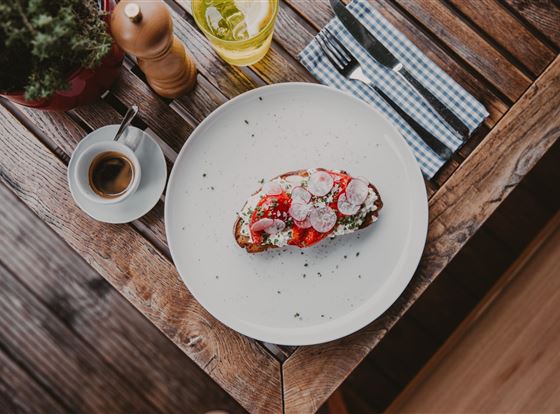 Ein Teller mit einem bunten Sandwich, belegt mit Tomaten und Radieschen, liegt auf einem Holztisch. Daneben befinden sich eine Tasse Kaffee und ein Glas mit einem erfrischenden Getränk.