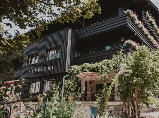Ein charmantes Gebäude mit der Aufschrift "SEEBICHL" umgeben von grünen Pflanzen. Die Fassade ist aus dunklem Holz und verleiht dem Ort eine gemütliche Atmosphäre.