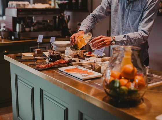 Ein Mann bereitet Getränke an einer Theke vor, die mit verschiedenen Köstlichkeiten und einer Obstschale dekoriert ist. Im Hintergrund sind Küchenutensilien und eine Kaffeemaschine zu sehen.