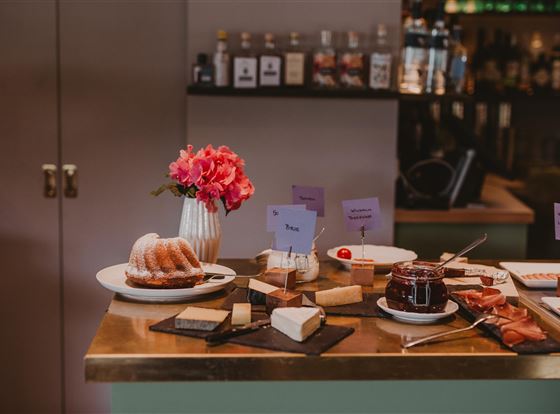 Ein Tisch mit verschiedenen Käsesorten, einem Gugelhupf und Marmelade. Blumen in einer Vase ergänzen die dekorative Anrichtung.