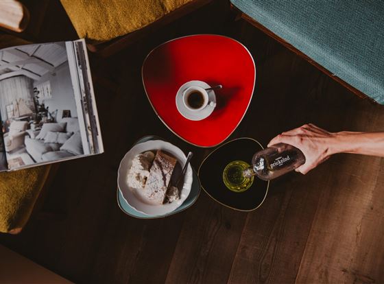Ein Tisch mit bunten Tellern, auf denen ein Stück Brot, ein Dessert und ein Getränk stehen. Dazu gibt es eine Tasse Kaffee und ein Magazin.