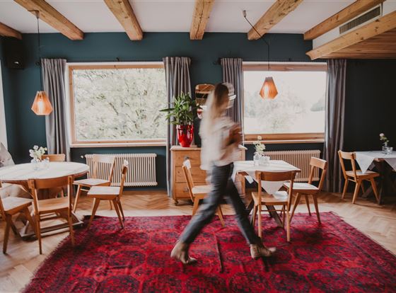 Ein modernes Restaurant mit Holzvertäfelung und großen Fenstern. Eine Person geht über einen roten Teppich zwischen den Tischen.