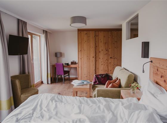 Ein modernes Hotelzimmer mit einem großen Bett und Holzelementen. Es gibt einen Schreibtisch, gemütliche Sitzgelegenheiten und große Fenster, die viel Licht hereinlassen.