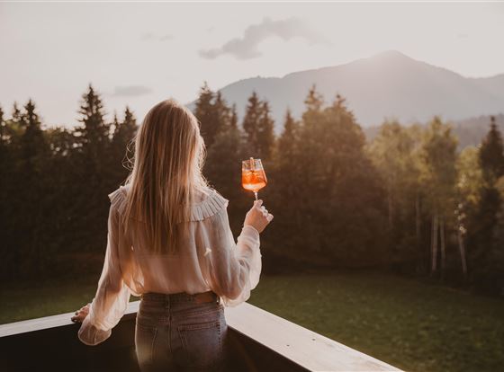 Eine Frau steht auf einem Balkon und hält ein Getränk in der Hand. Im Hintergrund sind grüne Wiesen und Berge bei Sonnenuntergang zu sehen.