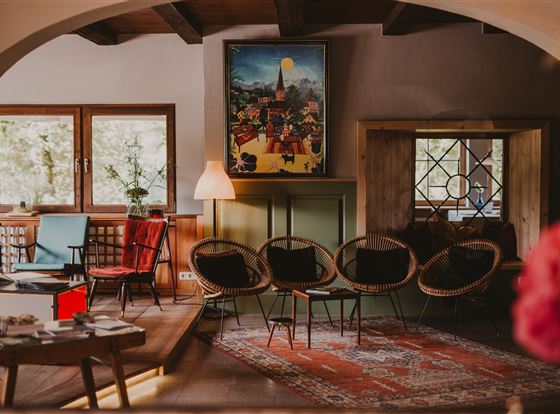 Ein gemütlicher Raum mit stilvollen Stühlen und einem bunten Gemälde an der Wand. Große Fenster lassen viel Tageslicht herein und schaffen eine einladende Atmosphäre.