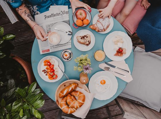 Ein Frühstückstisch mit frischen Croissants, Obst und Kaffee. Im Hintergrund liegt eine Zeitung und einige Personen sitzen entspannt.