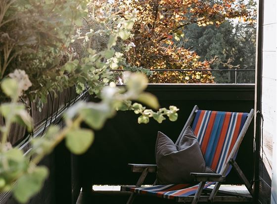 Eine gemütliche Terrasse mit einem bunten Liegestuhl und einem Kissen. Umgeben von Pflanzen und sanftem Licht.