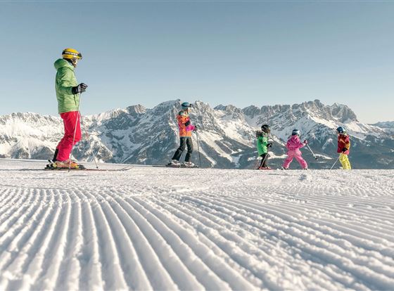 Eine Gruppe von Skifahrern auf einer schneebedeckten Piste. Im Hintergrund sind beeindruckende Berge sichtbar.