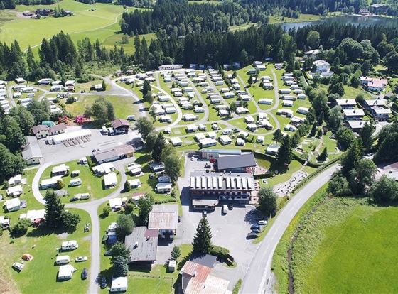 Ein großer Campingplatz mit vielen Wohnmobilen und Stellplätzen in einer grünen Landschaft. Im Hintergrund sind Wälder und sanfte Hügel zu sehen.