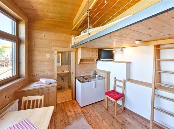 Ein gemütlicher Raum mit holzgetäfelten Wänden und einem Obergeschoss. Die Einrichtung umfasst eine Küche, einen Tisch und Stühle sowie eine Leiter zum oberen Bereich.