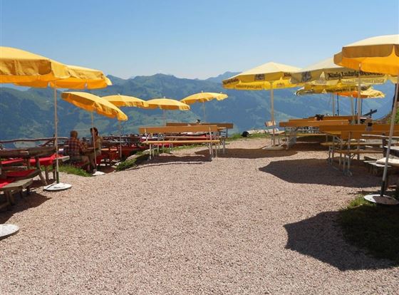 Ein schöner, sonniger Aussichtspunkt mit Tischen und Stühlen unter gelben Sonnenschirmen. Im Hintergrund erstrecken sich majestätische Berge.