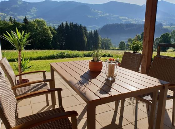 Eine gemütliche Terrasse mit einem Tisch und Stühlen, umgeben von üppiger Natur. Im Hintergrund sind sanfte Hügel und Bäume zu sehen.