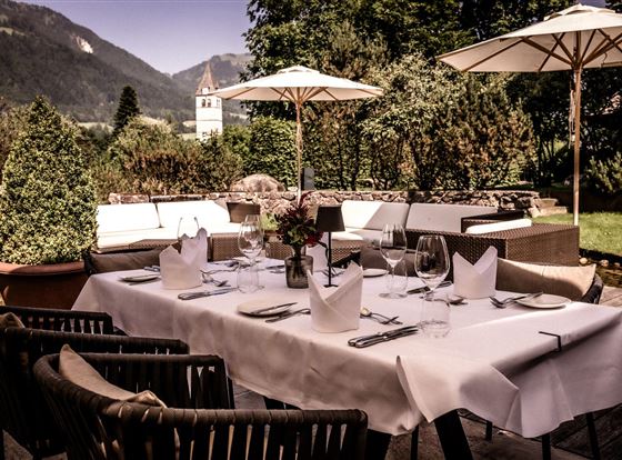 Ein elegantes Esszimmer im Freien mit einem dekorierten Tisch und Blick auf die Berge. Sonnenschirme und Pflanzen umgeben den Bereich und schaffen eine einladende Atmosphäre.