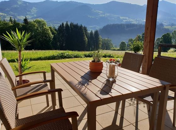 Ein schöner Außenbereich mit einem Tisch und Stühlen. Im Hintergrund sieht man grüne Hügel und Berge unter einem klaren Himmel.