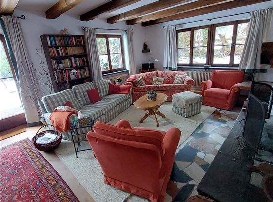 Ein gemütliches Wohnzimmer mit rustikalen Holzbalken und großen Fenstern. Es enthält ein rotes Sofa, einen runden Tisch und einen bunten Teppich.