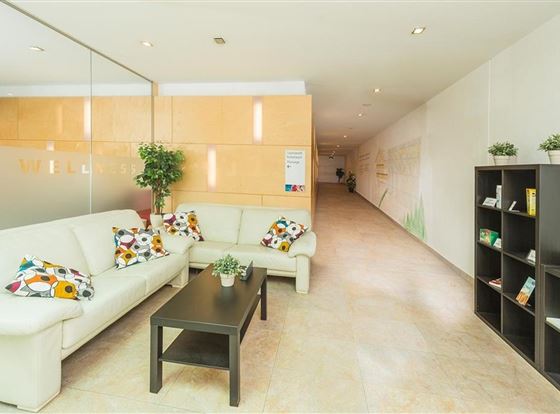 Ein modernes Wartezimmer mit einem weißen Sofa und einem kleinen Tisch. An der Wand sind Pflanzen und eine Bücherregal zu sehen.