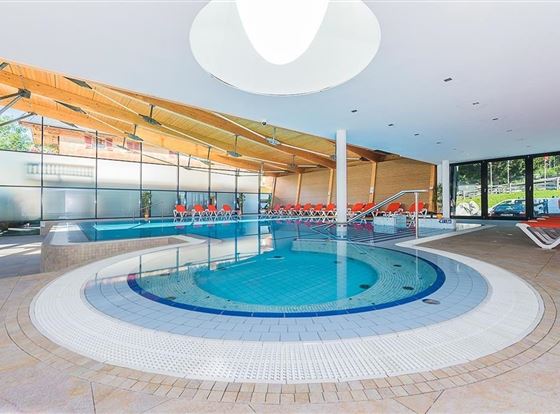 Ein moderner Innenpool mit einem runden Becken und einer hellen Holzdecke. Rote Liegen und große Fenster sorgen für eine einladende Atmosphäre.