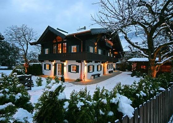 Ein gemütliches, traditionelles Haus im Winter, umgeben von Schnee und grünen Bäumen. Die Fenster sind warm beleuchtet, was eine einladende Atmosphäre schafft.