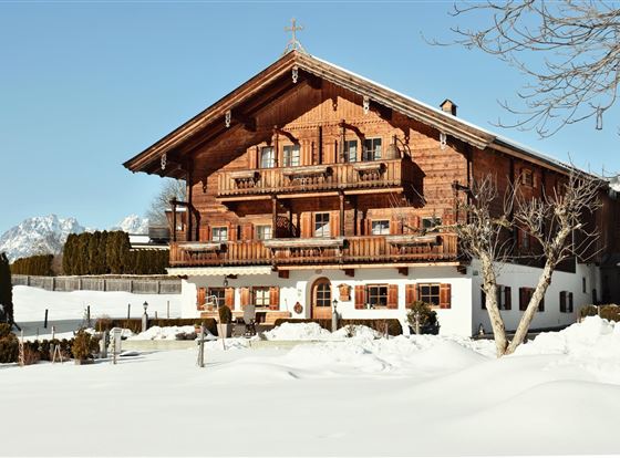Ein traditionelles alpenländisches Holzhaus in einer schneebedeckten Landschaft. Im Hintergrund sind Berge und ein klarer blauer Himmel sichtbar.