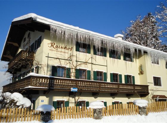 Ein charmantes Gebäude im Winter mit einem schneebedeckten Dach und Eiszapfen. Der klare blaue Himmel und die umgebende Landschaft verstärken die winterliche Atmosphäre.