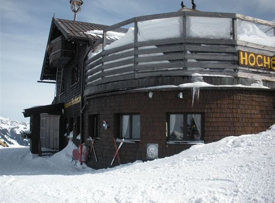 Ein gemütliches Berghaus, umgeben von Schnee. Auf der Terrasse sind Skiausrüstungen zu sehen.