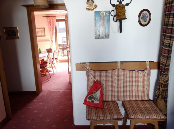 Ein gemütlicher Flur mit einem gepolsterten Sitzbereich und einer roten Tasche. Im Hintergrund ist ein helles Wohnzimmer mit einem Esstisch zu sehen.