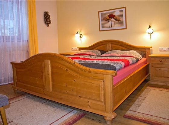 Ein gemütliches Schlafzimmer mit einem Holzbett und bunten Bettwäsche. Das Zimmer ist hell und freundlich gestaltet, mit einem Teppich und einem Fenster, das natürliches Licht hereinlässt.