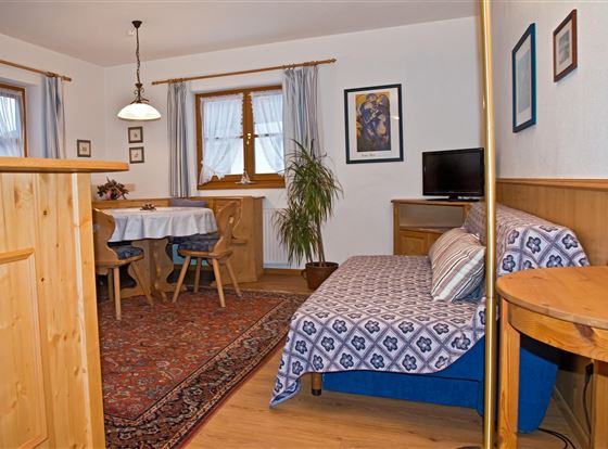 Ein gemütlicher Wohnraum mit einem Sofa, einem Tisch und Stühlen. Große Fenster lassen viel Tageslicht herein und der Raum ist hell und freundlich eingerichtet.