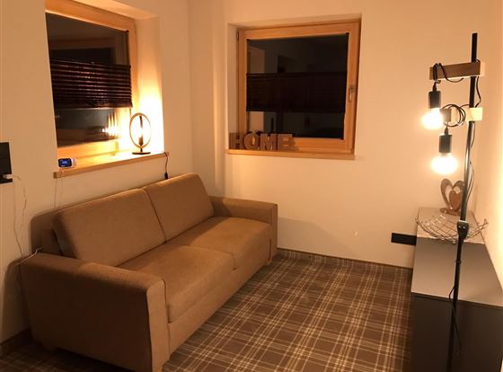 Ein gemütliches Wohnzimmer mit einem beigen Sofa und einem modernen Stehlampe. Es gibt zwei Fenster und die Wände sind hell gestrichen.