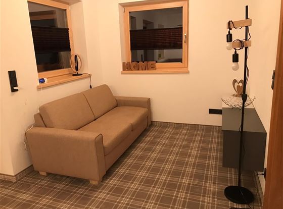 Ein gemütliches Wohnzimmer mit einem beigen Sofa und einem modernen Stehlampe. Die Wände sind hell und es gibt zwei Fenster mit Blick nach draußen.