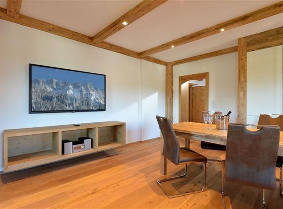 Ein modernes Wohnzimmer mit einem großen Fernseher an der Wand. Ein Holzst Tisch und elegante Stühle schaffen eine gemütliche Atmosphäre.