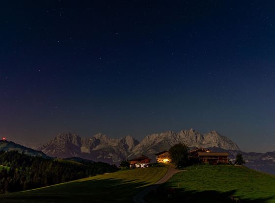 Eine ruhige Nachtlandschaft mit beleuchteten Hütten vor majestätischen Bergen. Der Himmel ist klar und voller Sterne.