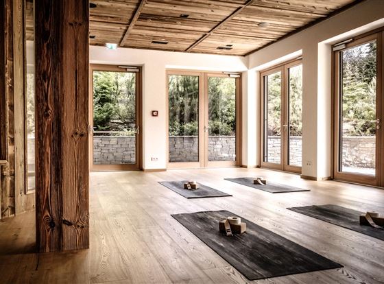 Ein heller Yogaraum mit Holzfußboden und großen Fenstern. Auf dem Boden liegen Yogamatten und Blöcke, um eine entspannte Atmosphäre zu schaffen.