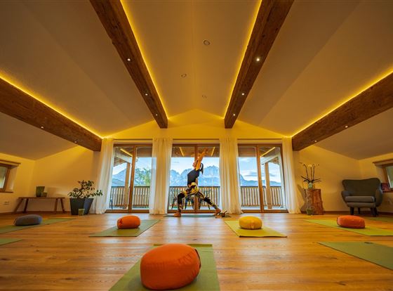 Ein heller und einladender Yogaraum mit großen Fenstern und Holzbalken. Auf dem Boden liegen Yogamatten und ein paar orangefarbene Sitzkissen.