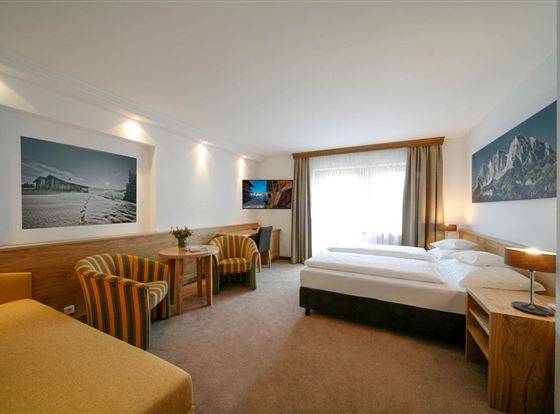 Ein modernes Hotelzimmer mit zwei Einzelbetten, einem gemütlichen Sitzbereich und großen Fenstern. Die Wände sind mit Bildern der Berge dekoriert, und es gibt einen Fernseher im Zimmer.