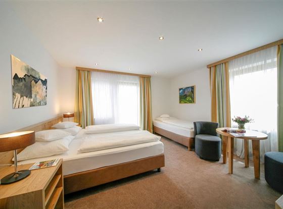 Ein helles und modernes Zimmer mit zwei Betten und großen Fenstern. Es gibt gemütliche Sitzgelegenheiten und dekorative Bilder an den Wänden.