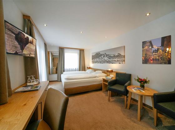 Ein modernes Hotelzimmer mit einem Doppelbett und zwei Sessel. Der Raum ist hell und freundlich eingerichtet, mit Bildern an den Wänden.