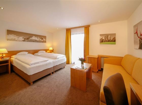 Ein gemütliches Hotelzimmer mit zwei Einzelbetten und einem Sofa. Große Fenster lassen viel Tageslicht herein und sorgen für eine freundliche Atmosphäre.