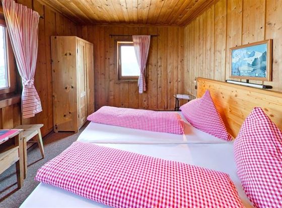 Ein gemütliches Zimmer mit Holzvertäfelung und großen Fenstern. Es sind zwei Betten mit rot-weiß karierten Kissen und eine einfache Einrichtung vorhanden.