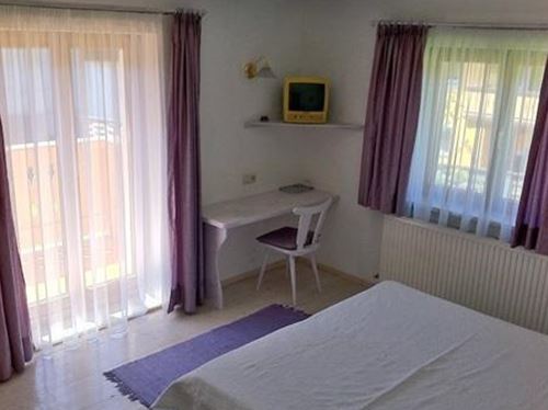 Ein helles Schlafzimmer mit einem großen Bett und einem kleinen Schreibtisch. Große Fenster mit Vorhängen lassen viel Licht herein.