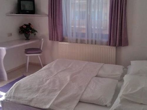 Ein einfaches Schlafzimmer mit einem großen Bett und weißen Bettwäsche. Der Raum hat lila Vorhänge und einen kleinen Schreibtisch.