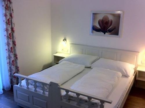 Ein helles Schlafzimmer mit einem gemütlichen Doppelbett und weißen Bettwäsche. An der Wand hängt ein Bild von einer Blume, und das Fenster hat gardinen mit Blumenmuster.