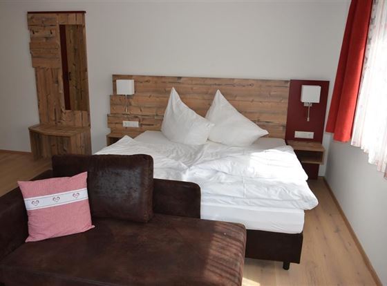 Ein gemütliches Schlafzimmer mit einem Doppelbett und rustikalem Holzdesign. Neben dem Bett steht ein dunkles Sofa mit einem dekorativen Kissen.
