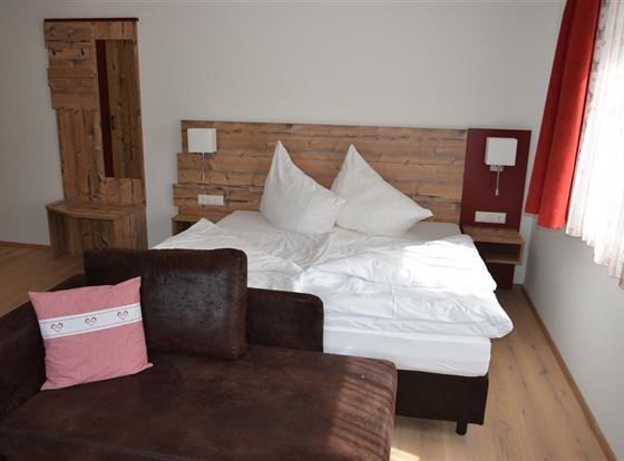 Ein gemütliches Schlafzimmer mit einem Bett, weißen Kissen und einem dunklen Sofa. Helle Wände und Fenster lassen viel Licht herein.