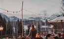 Eine Frau steht vor einem Feuerscheit auf einer Terrasse mit Blick auf die Berge. Lichterketten und eine entspannte Atmosphäre schaffen eine gemütliche Stimmung.