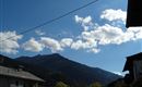 Ein klarer Himmel mit einigen Wolken über den Bergen. Im Vordergrund sind Häuser und Bäume zu sehen.