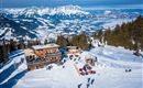 Eine malerische Berglandschaft mit schneebedeckten Pisten und einer gemütlichen Hütte. Im Hintergrund erstrecken sich majestätische Berge unter einem klaren blauen Himmel.