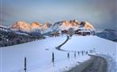 Eine schneebedeckte Landschaft mit malerischen Bergen im Hintergrund. Kleine Holzhäuser und ein verschneiter Weg führen durch die winterliche Szene.
