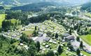 Ein großer Campingplatz in einer grünen Umgebung mit vielen Wohnmobilen und Stellplätzen. Im Hintergrund sieht man sanfte Hügel und einen kleinen See.